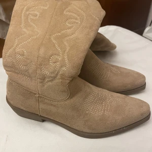 Helt nya Beiga Cowboyboots/stövlar  - Helt nya snygga beiga cowboyboots/stövlar med dekorativa sömmar och dragkedja på sidan. Bootsens klassiska westernstil och spetsiga tå ger en trendig look. Perfekta för dig som vill sticka ut med en cool och avslappnad stil.                                                                       Maxhöjd: 39 cm Bredd på skaft som bredast: 19 cm 
