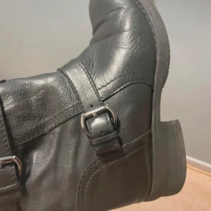 Svarta boots med spännen och dragkedja - Snygga svarta boots i skinn med tre dekorativa spännen i silverfärg och dragkedja på insidan för enkel påtagning. Klassisk rund tå och platt sula, perfekt till jeans eller kjol. Bootsens stilrena design gör dem lätta att matcha.
