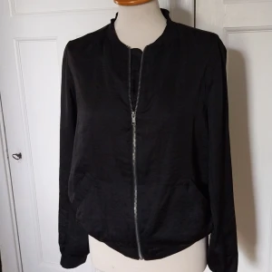 Svart bomberjacka från Åhléns, stl 40 - Svart bomberjacka från Åhléns i storlek 40. Jackan har klassisk rundad krage, dragkedja framtill och två snedställda fickor. Tillverkad i ett lätt tyg med ribbade muddar vid hals och ärmslut. Perfekt för en clean och stilren look.