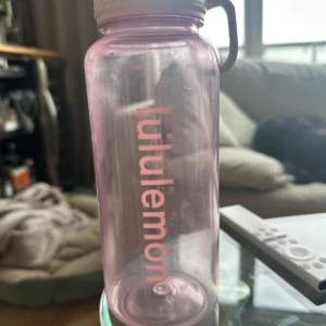 Lululemon vattenflaska - Rosa vattenflaska från Lululemon med 900 ml kapacitet. Flaskan har tydliga volymmärkningar och ett praktiskt bärhandtag. Den är använd men är i gott skick men finns tecken på slitage. Perfekt för träning, gym eller vardagsbruk.