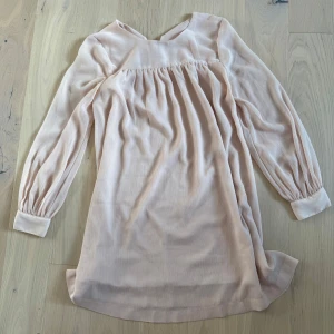 Ljusrosa kort klänning med långa ärmar - Superfin ljusrosa klänning med långa ärmar och breda manschetter med knappar. Klänningen har en lös passform, lätt rynk vid bysten och dragkedja i ryggen. Perfekt för dig som gillar en enkel men söt stil.