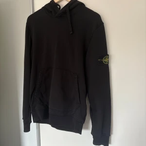 Svart hoodie från Stone Island - Svart hoodie från Stone Island med klassisk patch på vänster ärm. Tröjan har huva med snörning, magficka och ribbade muddar. Inköpt på Nk för 3500kr. Har tyvärr inte kvitto men går att legit checka via certi logo. Har inte tvättats mer än 4 gånger så suveränt skick! 