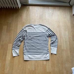 Randig långärmad tröja från moncler  - Säljer en klassisk vit långärmad tröja från moncler med svarta horisontella ränder. Tröjan har rund halsringning och en liten logotyp på ärmen. Perfekt för en avslappnad och stilren look. Obs! Har tre små hål framtill!