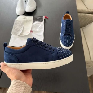 Blå Christian Louboutin sneakers med nitar - Säljer ett par blå sneakers från Christian Louboutin i mocka med coola nitar på tån och klassisk röd sula. Skorna har snörning och vit platt sula, perfekt för dig som vill sticka ut med en lyxig och edgy stil.