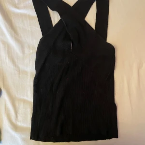 Svart ribbad korsett-topp - Säljer en svart ribbad korsett-topp med breda axelband som korsas i ryggen. Toppen är ärmlös och har en tight passform som framhäver figuren. Perfekt för att skapa en cool och trendig look.