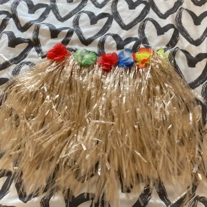 Beige hawaiikjol med färgglada blommor - Säljer en beige hawaiikjol i syntetmaterial med klassiska plaststrån och färgglada blommor i midjan. Kjolen har kardborreknäppning i midjan för enkel på- och avtagning. Perfekt för temafest eller sommarhäng! Storleken är justerbar 