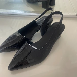 Svarta pumps med spetsig tå och klack - Snygga svarta pumps med spetsig tå och låg klack. Skorna har en croco-präglad yta i glansigt syntetmaterial och öppen häl med rem. Perfekta för dig som vill ha en stilren och trendig look.