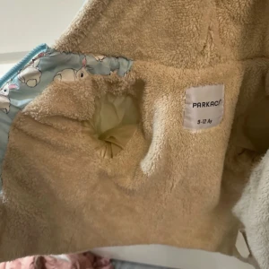 Blå teddyjacka med kaninmönster Parkaci - Supersöt blå teddyjacka från Parkaci med gulligt kaninmönster och mysigt beige teddyfoder. Jackan har huva och dragkedja framtill, perfekt för kyliga dagar. Insidan är mjuk och varm, utsidan har ett lekfullt print som sticker ut.