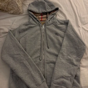 Grå hoodie från Burberry med dragkedja - Priset kan prutas, det är bara att skruva. Säljer en grå hoodie från Burberry med klassiskt rutigt foder i huvan. Tröjan har dragkedja framtill, snörning vid huvan och diskret Burberry-logga på bröstet. Perfekt för dig som gillar stilrena och exklusiva plagg.