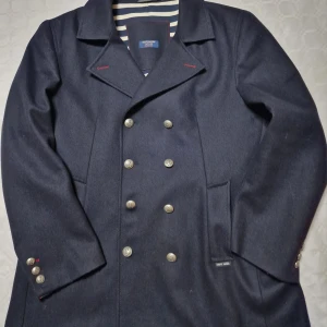 Marinblå skepparkavaj från Saint James - Snygg marinblå skepparkavaj från Saint James med klassisk dubbelknäppning och silvriga knappar med ankardetaljer. Jackan har bred krage, randigt innerfoder i blått och vitt samt röda sömdetaljer vid kragen. Perfekt för dig som gillar tidlös och nautisk stil.