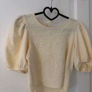 Beige puffärm blus från Monki - Supersöt beige/ gul  blus från Monki med puffiga korta ärmar och strukturmönster i tyget. Toppen har rund halsringning, öppen rygg med knyt och elastiska ärmslut. 