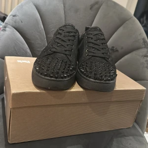 Svarta Christian Louboutin sneakers med nitar - Säljer ett par svarta sneakers från Christian Louboutin i mocka med coola svarta nitar på tån och klassisk röd sula. Skorna har snörning och en låg profil, perfekt för dig som vill sticka ut med en edgy vibe. Storlek 42 men passar 43