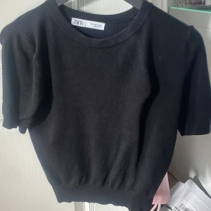 Svart stickad tröja från Zara - En svart stickad tröja från Zara med rund halsringning och korta ärmar. Tröjan har en enkel och stilren design med ribbad nederkant som ger en snygg passform. Perfekt att matcha med jeans eller kjol för en clean look.