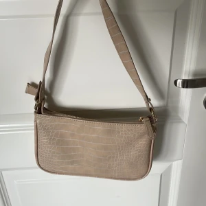 Beige handväska  - Snygg beige axelväska med guldiga detaljer och dragkedja. Väskan kommer med två olika axelremmar för att kunna variera stilen. Perfekt storlek, finns även ett mindre fack i väskan. Denna väskan är i mycket bra skick då den bara har använts ett fåtal gånger.