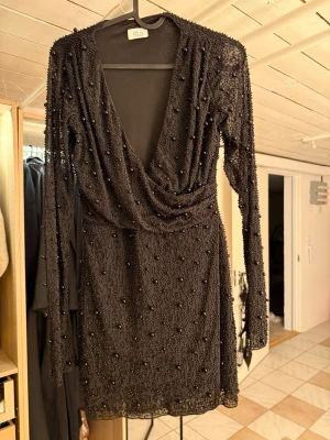 Svart glittrig kortklänning Ivyrevel - Supersnygg svart kortklänning från Ivyrevel med djup v-ringning och långa ärmar. Klänningen är täckt av små pärlor och paljetter som ger ett glittrigt intryck. Perfekt för dig som vill sticka ut! Använd 1 gång.
