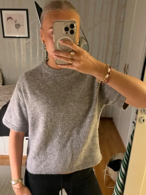 Grå stickad tröja från Gina Tricot - Mysig grå stickad tröja från Gina Tricot i ullblandning. Tröjan har korta ärmar, rund halsringning och en rak, lite boxig passform. Perfekt att slänga på sig över ett linne eller t-shirt för en chill look. Aldrig använd!