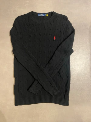 Svart kabelstickad tröja Polo Ralph Lauren - Klassisk svart kabelstickad tröja från Polo Ralph Lauren med röd broderad logga på bröstet. Rund halsringning och ribbade muddar vid ärmslut och nederkant. Perfekt till lager på lager under höst och vinter.