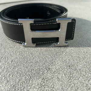 Svart Bälte - Snyggt svart bälte med ikoniskt H-spänne i silverfärgad metall. Bältet är gjort i äkta skinn med vit kontrastsöm längs kanterna. Perfekt accessoar för att lyfta din outfit med en lyxig touch.