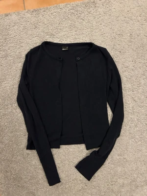 Marinblå  kofta från Gina Tricot - En enkel och stilren marinblå kofta från Gina Tricot. Modellen är rak med rund halsringning och långa ärmar. Koftan har en knapp upptill och är tillverkad i mjukt, tunt stickat material. Perfekt att slänga över en topp eller klänning.