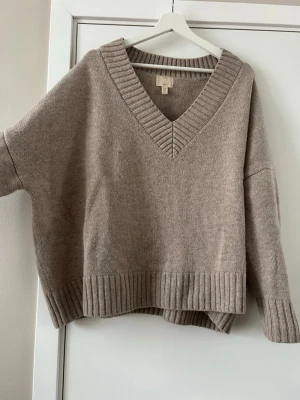 Beige stickad tröja från H&M - Mysig beige stickad tröja från H&M i 100% ull. Tröjan har en djup v-ringning, bred ribbad kant vid hals, ärmslut och nederkant samt en loose passform. Perfekt för lager på lager under höst och vinter.