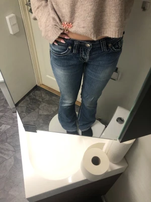 Blå bootcut jeans med kontrastsömmar - Säljer ett par blå bootcut jeans från AMO med tydliga kontrastsömmar och snygga detaljer på fickorna. Lågmidjade och med klassisk femficksdesign. Jeansen har slitningar framtill och bak, samt dekorativa knappar och dragkedja.