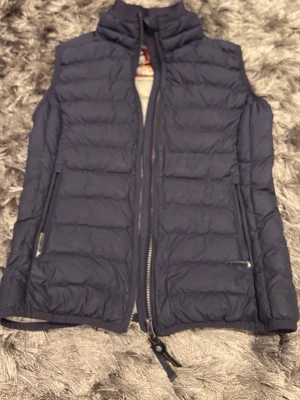 Mörkblå dunväst från Parajumpers S äkta - Säljer en mörkblå dunväst från Parajumpers med quiltad design och dragkedja framtill. Västen är superlätt och har två fickor med dragkedja. Insidan är ljusgrå och har flera snygga märkespatchar. Perfekt för lager-på-lager under kyliga dagar. Pris kan diskuteras!
