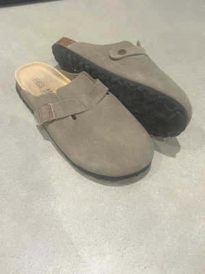Beige mockasandaler från Birkenstock - Säljer ett par klassiska Birkenstock sandaler i beige mocka med justerbart spänne i metall. Skorna har en mjuk innersula och svart räfflad yttersula för extra komfort. Perfekta för dig som gillar stilren och avslappnad look.