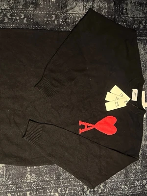 Svart stickad tröja från Ami - Svart stickad tröja från Ami med ikonisk röd hjärtlogga och bokstaven A på bröstet. Tröjan har rund hals, ribbade muddar och är långärmad. Perfekt för dig som vill ha en clean men ändå statement look.