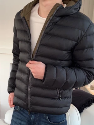Svart pufferjacka från Colmar - Säljer en svart pufferjacka från Colmar Originals med huva och dragkedja. Jackan har en quiltad design och två fickor med dragkedja. Insidan är olivgrön och materialet är syntet. Perfekt för kalla dagar och riktigt snygg till jeans.