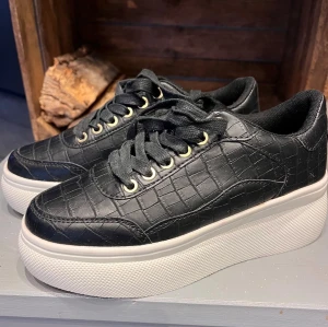 Svarta sneakers med platåsula från Duffy - Snygga svarta sneakers från Duffy med croco-mönstrad yta och vita platåsulor. Skorna har guldfärgade öljetter, svarta snören och en rund tå. Materialet är syntet med textilfoder. though vibes till. Perfekta för dig som vill ha lite extra höjd.