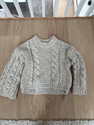 Beige stickad tröja från NA-KD - Supermysig beige stickad tröja från NA-KD med grova flätstickade detaljer på både ärmar och framstycke. Tröjan har en ribbad kant vid hals, ärmslut och nederkant. Perfekt för kyliga dagar och ger en chill vibe till din outfit.