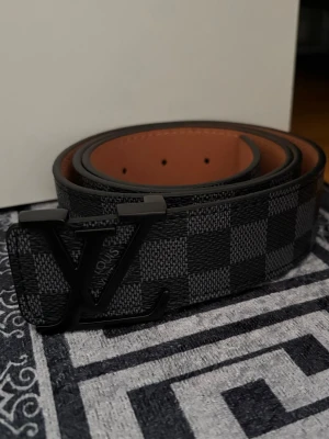L V bälte 110cm  - Säljer ett svart och grått bälte med klassiskt rutigt mönster. Bältet har en lyxig känsla och insidan är brun. Perfekt accessoar för att lyfta din outfit.