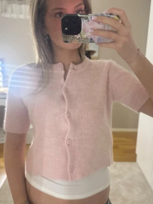 Ljusrosa stickad kofta med kort ärm - Superfin ljusrosa stickad kofta med korta ärmar och rund hals. Koftan är croppad och har knappar framtill. Perfekt att slänga över ett linne för en söt och chill look. Ribbstickad och mjuk i materialet.