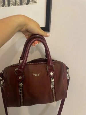 Vinröd handväska Zadig & Voltaire - Säljer en vinröd handväska från Zadig & Voltaire i skinn. Väskan har två rejäla handtag, avtagbar axelrem och snygga silverfärgade dragkedjor framtill. Klassisk boxig form och diskret logga med vingar på framsidan. Perfekt storlek för det viktigaste.