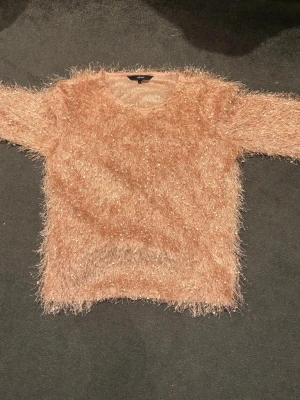 Glittrig fluffig tröja från Vero Moda - Supersöt fluffig tröja från Vero Moda i ljusrosa med glittriga inslag. Tröjan har lång ärm och är stickad med ett lurvigt material som ger en mysig och trendig look. Perfekt för dig som vill sticka ut lite extra!