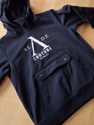 Svart skidjacka från Slade Couture - Snygg svart skidjacka från Slade Couture med stor logga framtill och mindre logga bak. Jackan har huva, stor ficka med knappstängning på magen och ribbade muddar. Perfekt för skidåkning.