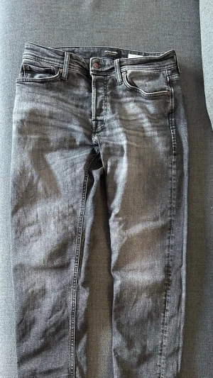 Grå jeans från Jack & Jones - Säljer ett par grå jeans från Jack & Jones med klassisk femficksdesign och smal passform. Jeansen har en snygg tvättad look och är tillverkade i mjukt bomullsmaterial med lite stretch för extra komfort. Perfekta för en avslappnad stil. Storlek 29/30 modell taperd Mike.