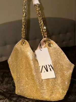 Glittrig guld axelväska från Zara - Säljer en superglittrig guld axelväska från Zara med kedjehandtag i metall. Väskan har en mjuk, flexibel form och är täckt av små glittrande stenar som verkligen fångar ljuset. Perfekt för dig som vill sticka ut och addera lite extra bling till din outfit.