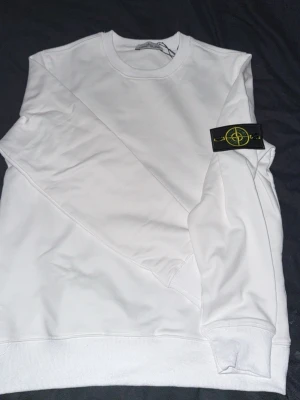 Vit sweatshirt från Stone Island - Säljer en clean vit sweatshirt från Stone Island med den klassiska svarta loggan på vänster ärm. Tröjan har rund hals, långa ärmar och ribbade muddar. Tillverkad i mjuk bomull för skön känsla och enkel stil.