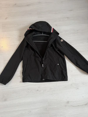 Svart vindjacka från Moncler - Snygg svart vindjacka från Moncler med huva och dragkedja framtill. Jackan har Moncler-logga på ärmen och coola ränder i rött, vitt och blått på huvan. Tillverkad i lätt polyester, perfekt för blåsiga dagar. Två fickor med tryckknappar och elastiska muddar vid ärmslut. En av tryckknapparna har lossnat så finns endast på ena sidan, såklart dras priset ner på grund av detta. Inget bestämt pris så bara höra av sig!