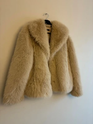 Beige fluffig pälsjacka - Säljer en supermjuk och fluffig beige pälsjacka med bred krage. Jackan har en oversized look och är perfekt för dig som vill sticka ut med en trendig och varm stil. Passar till alla tillfällen när du vill känna dig extra cozy och snygg.