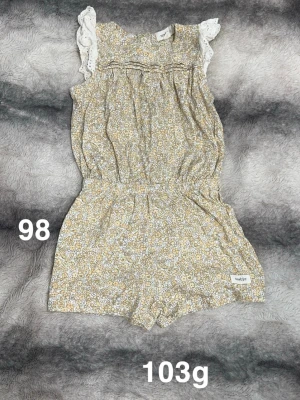 Blommig byxdress från Newbie - Söt blommig byxdress från Newbie i ljusa toner. Byxdressen har korta ben, resår i midjan och volangdetaljer vid axlarna i spets. Perfekt för varma dagar och riktigt bekväm att bära.🌸 Fint använt skick. Storlek 98 