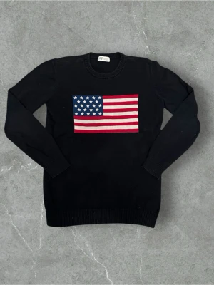 Marinblå stickad tröja med USA-flagga H&M - Marinblå stickad tröja från H&M med stor amerikansk flagga på bröstet. Klassisk rund halsringning och ribbade muddar vid ärmslut och nederkant. Perfekt för dig som vill sticka ut med en cool detalj.