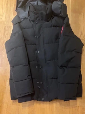 Svart dunjacka från Canada Goose - Säljer en svart dunjacka från Canada Goose med huva och klassisk patch på ärmen. Jackan har quiltad design, långa ärmar och knäppning med både dragkedja och knappar. Perfekt för kalla dagar och riktigt snygg till vintern.