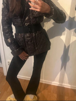 Quiltad Moncler pufferjacka brun - Säljer en mörkbrun quiltad pufferjacka från Moncler med bälte i midjan och dragkedja. Jackan har lång ärm och ikonisk Moncler-logga på ärmen. Materialet är glansigt och känns riktigt lyxigt, perfekt för kalla dagar.