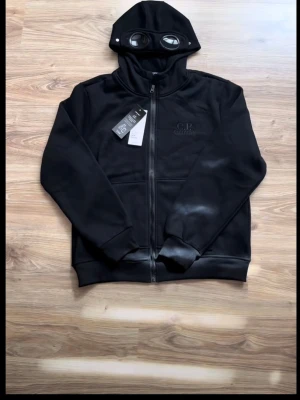 C.P Company - C.P. Company goggle hoodie - ny med tags  Säljer en svart zip hoodie från C.P. Company med den ikoniska goggle-hooden. Riktigt clean och eftertraktad modell inom streetwear.