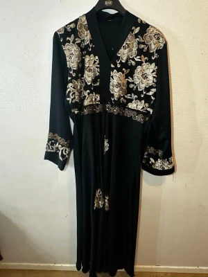 Svart långklänning med guldbroderi - Svart långklänning med lyxiga guldfärgade blommiga broderier på axlar, bröst och ärmslut. Klänningen har lång ärm och ett elegant fall. Perfekt för dig som vill sticka ut med detaljer i guld och blommönster.