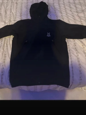 Svart vindjacka från Haglöfs - Säljer en svart vindjacka från Haglöfs med huva och två praktiska fickor med dragkedja. Jackan har minimalistisk design och är tillverkad i slitstarkt syntetmaterial. Perfekt för dig som vill ha en clean och funktionell look.
