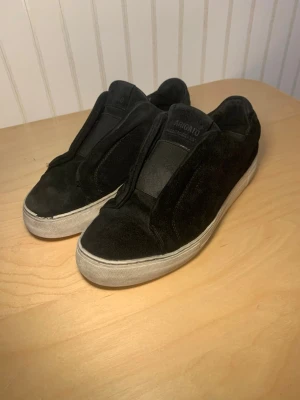  Axel Arigato skor - Säljer ett par svarta sneakers från Axel Arigato i mjuk mocka. Skorna har en stilren design utan snörning, med vit platt sula och diskret logga på sidan. Perfekta för dig som gillar minimalistisk och clean stil. Defekt hål i sulan. 