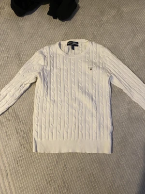 Vit kabelstickad tröja från GANT - Kabelstickad vit tröja från GANT med rund halsringning och diskret logga på bröstet. Tröjan har lång ärm och ribbade muddar vid ärmslut och nederkant. Perfekt för dig som gillar klassisk och stilren look.